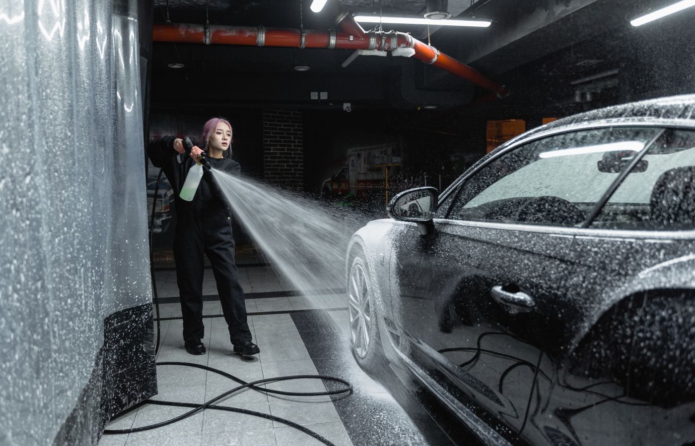 Person using a high-pressure wand to rinse soap off a dark car inside a self-service wash bay