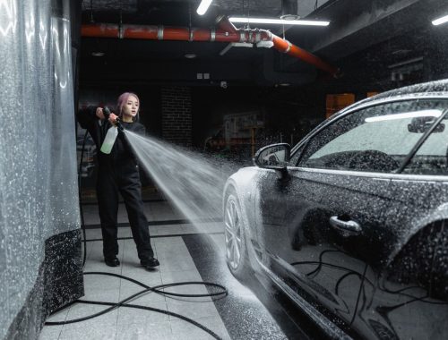 Person using a high-pressure wand to rinse soap off a dark car inside a self-service wash bay