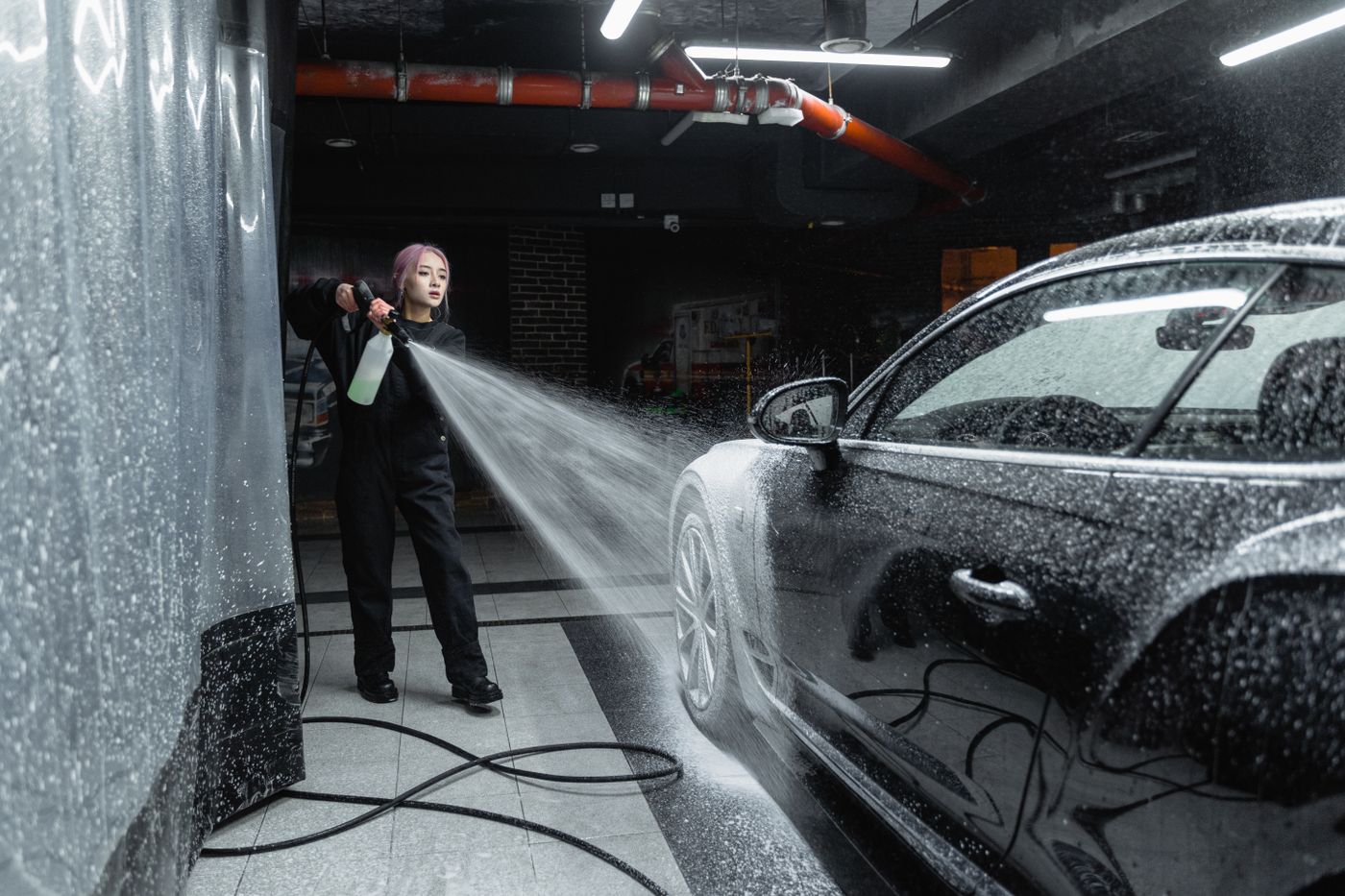 Person using a high-pressure wand to rinse soap off a dark car inside a self-service wash bay